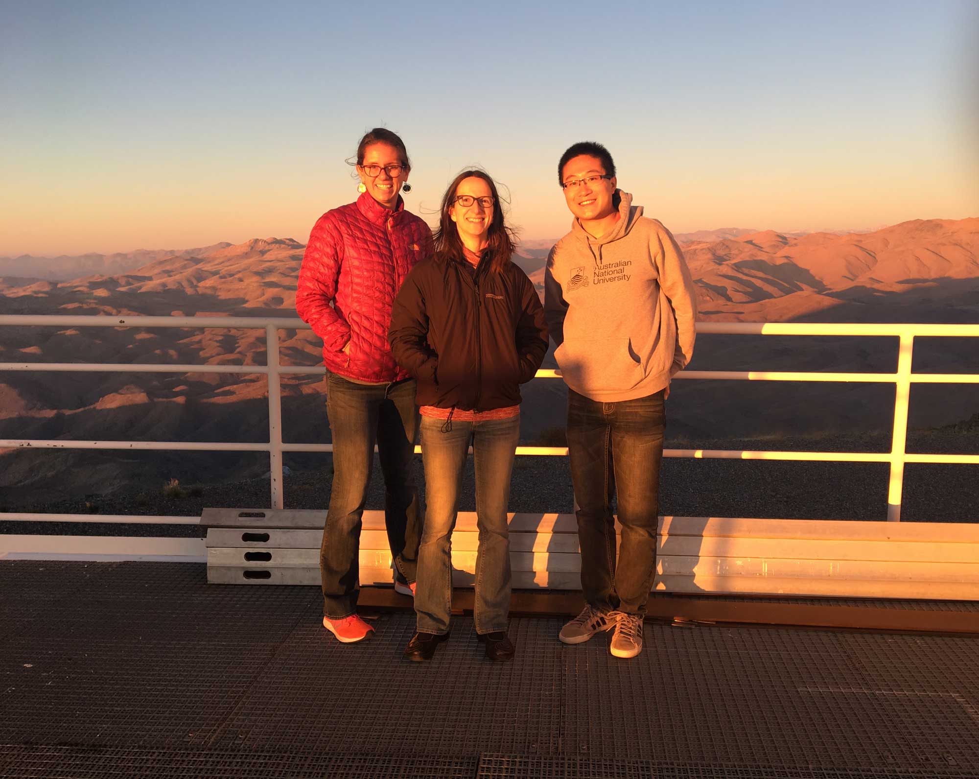 Three people stand on an overlook with mountains beyond.