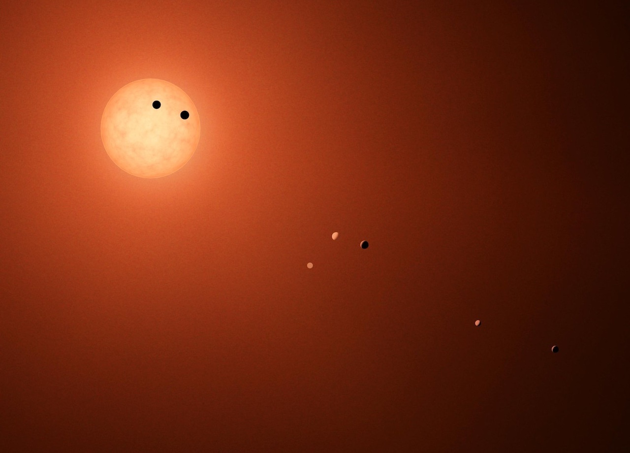 Artist's concept shows the red-dwarf star, TRAPPIST-1, at the upper left, with two large dots on the face of the disk representing transiting planets; five more planets are shown at varying positions descending toward the lower right as they orbit the star.